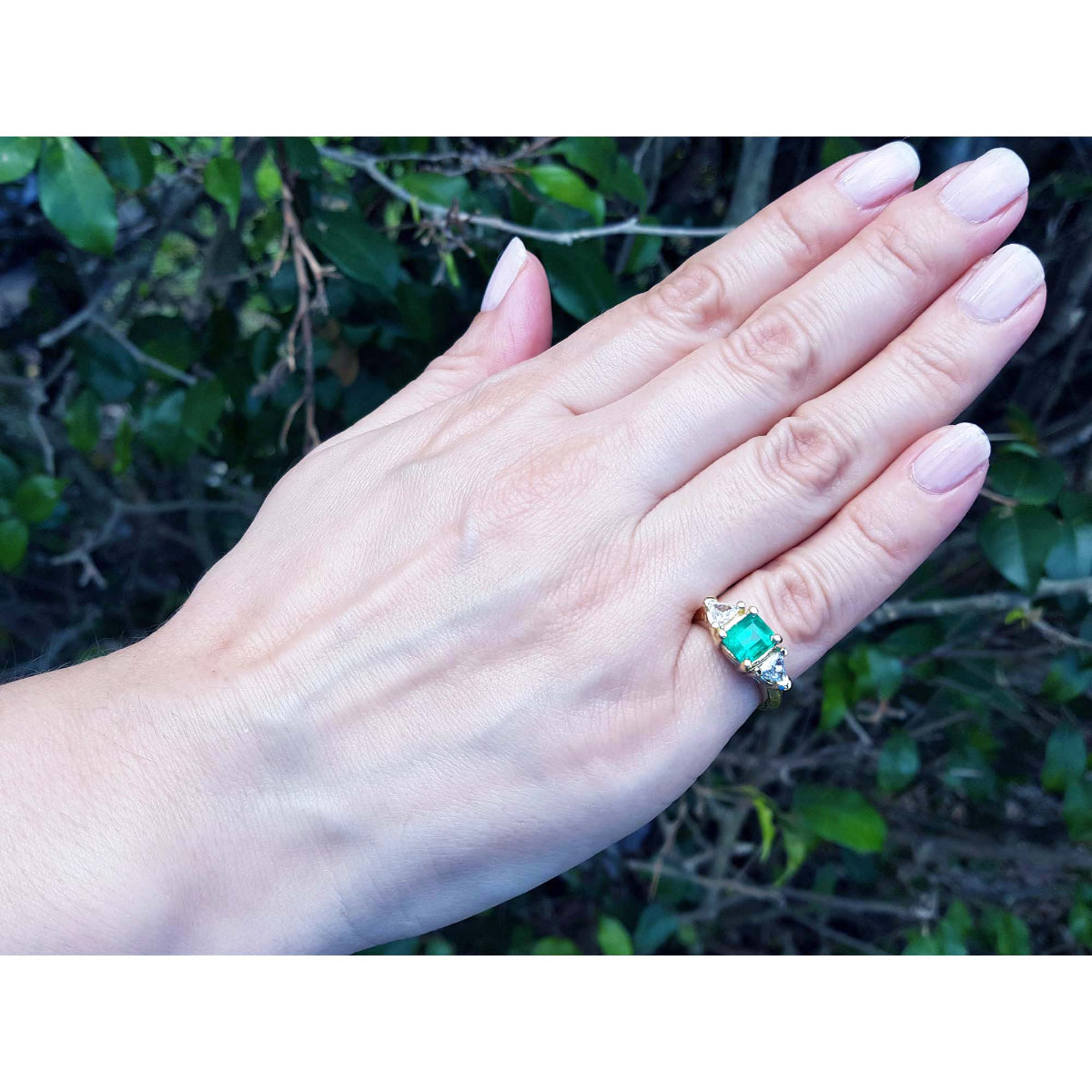 Hand wearing a gold ring with a natural colombian emerald and trillion diamonds against a natural background.