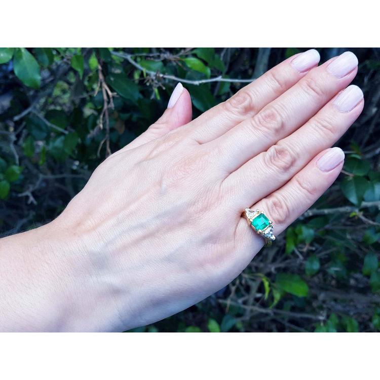 Hand wearing a gold ring with a natural colombian emerald and trillion diamonds against a natural background.