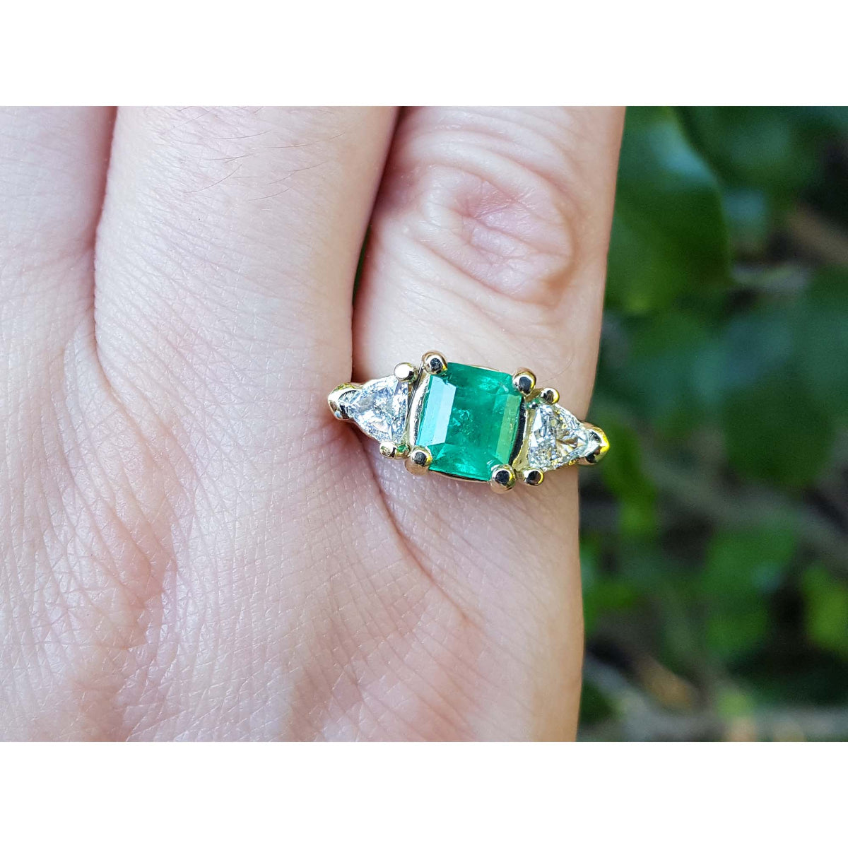 A 18k yellow gold genuine Muzo colombian emerald ring with trillion diamonds on a finger against a blurred natural background.