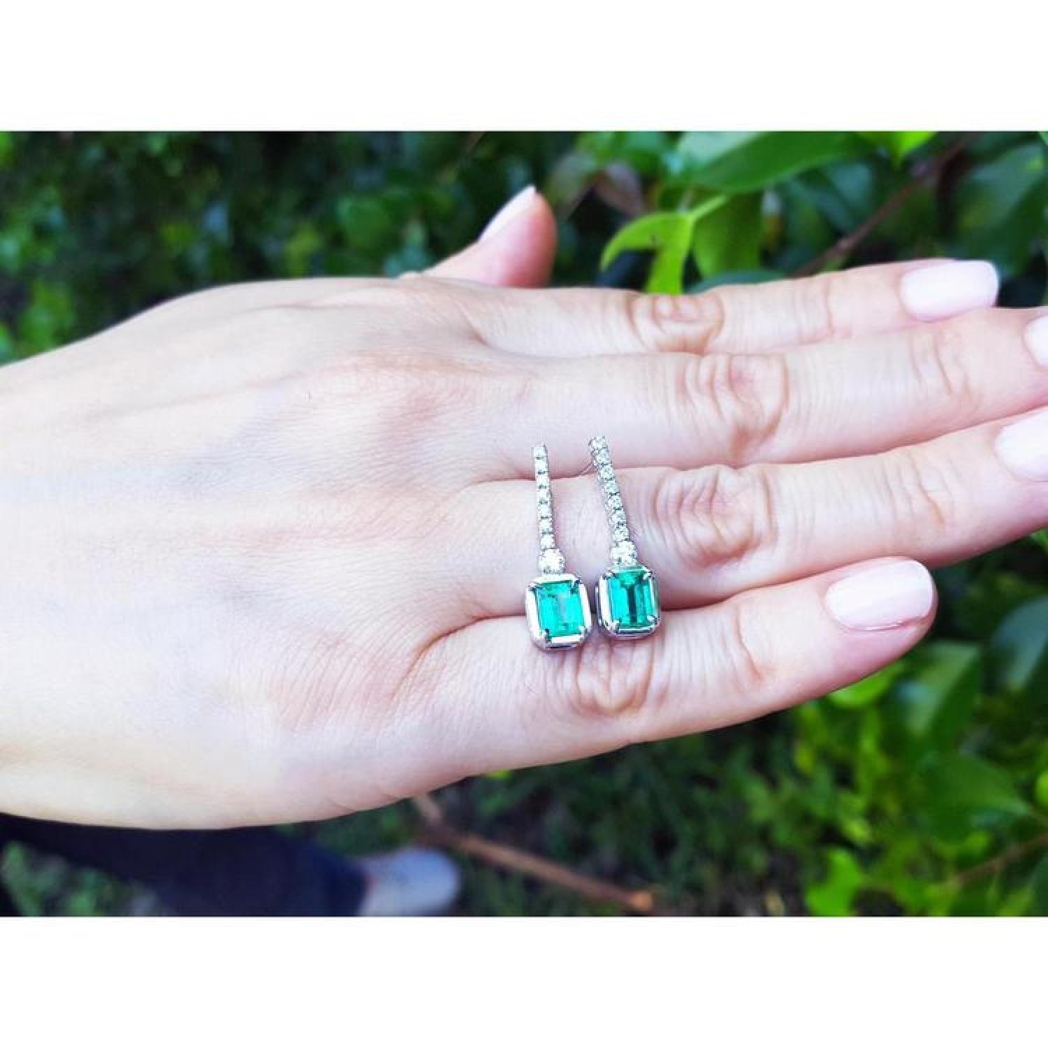 18K white gold dangling emerald-cut emerald earrings with diamond-studded lines on a hand.