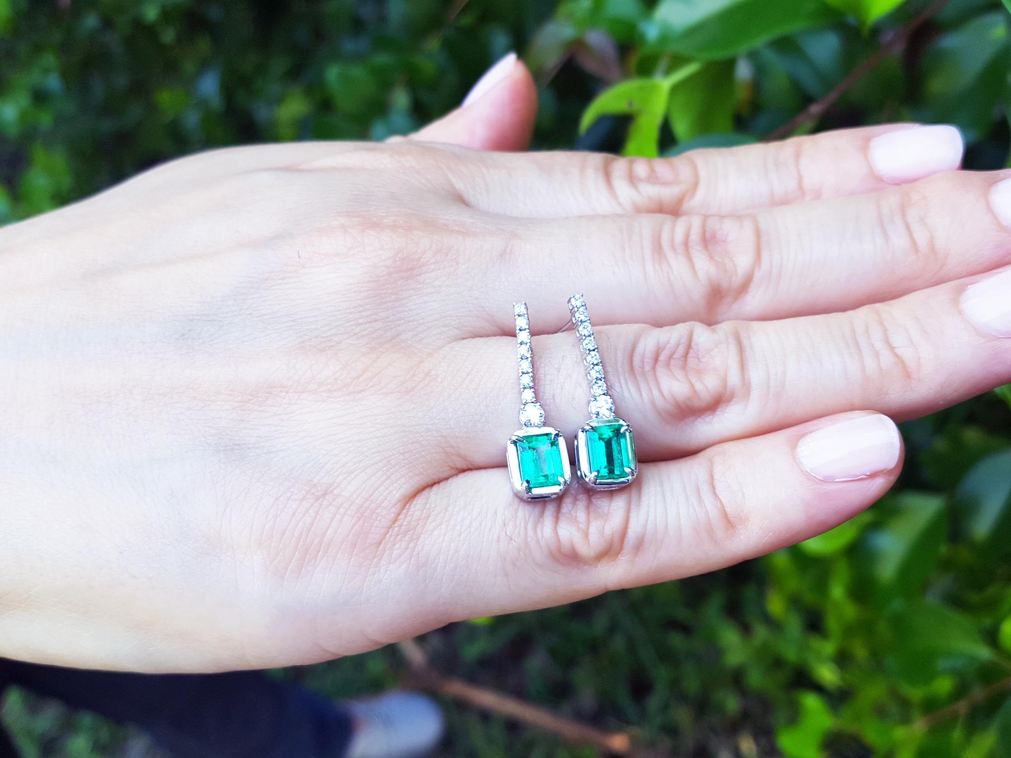 A pair of emerald dangle earrings placed on a hand with a blurred green leafy background