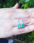 A pair of emerald dangle earrings placed on a hand with a blurred green leafy background