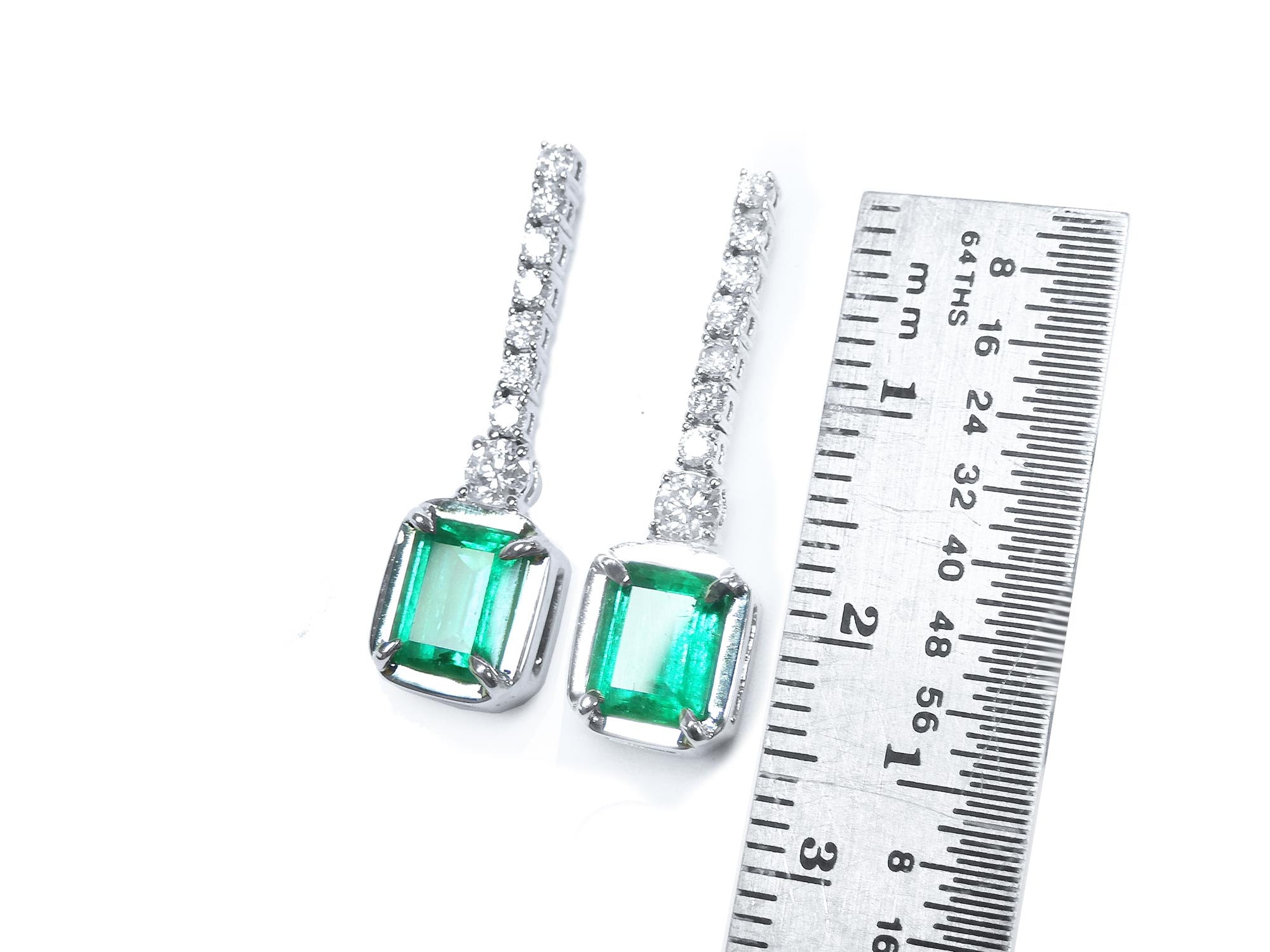 Pair of 18k white gold emerald dangle earrings next to a ruler for scale on a white background.