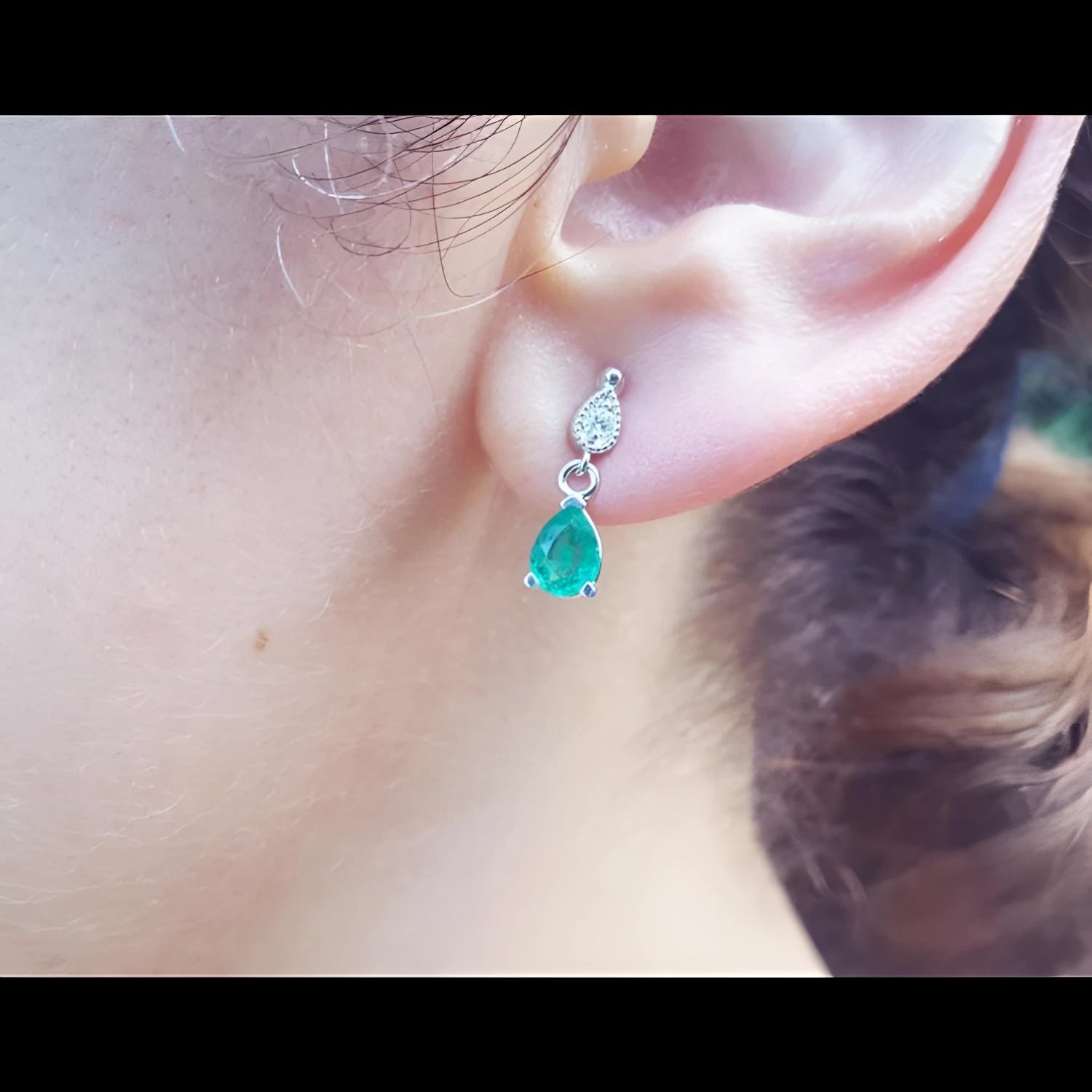 Dangling teardrop emerald and diamond earrings in 14K white gold on an ear.