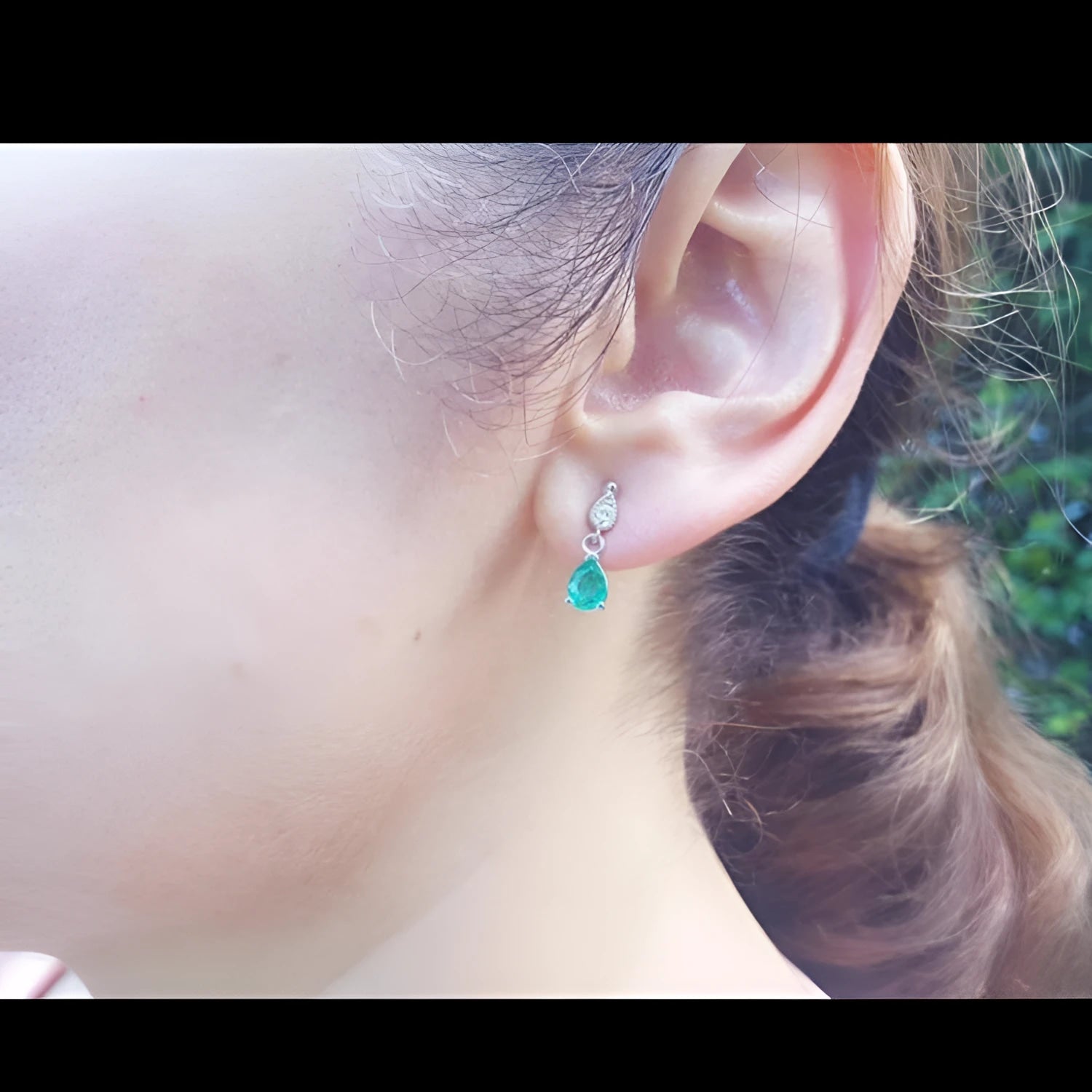 Dangling teardrop emerald earrings in 14K white gold with diamond accents on a woman's ear.