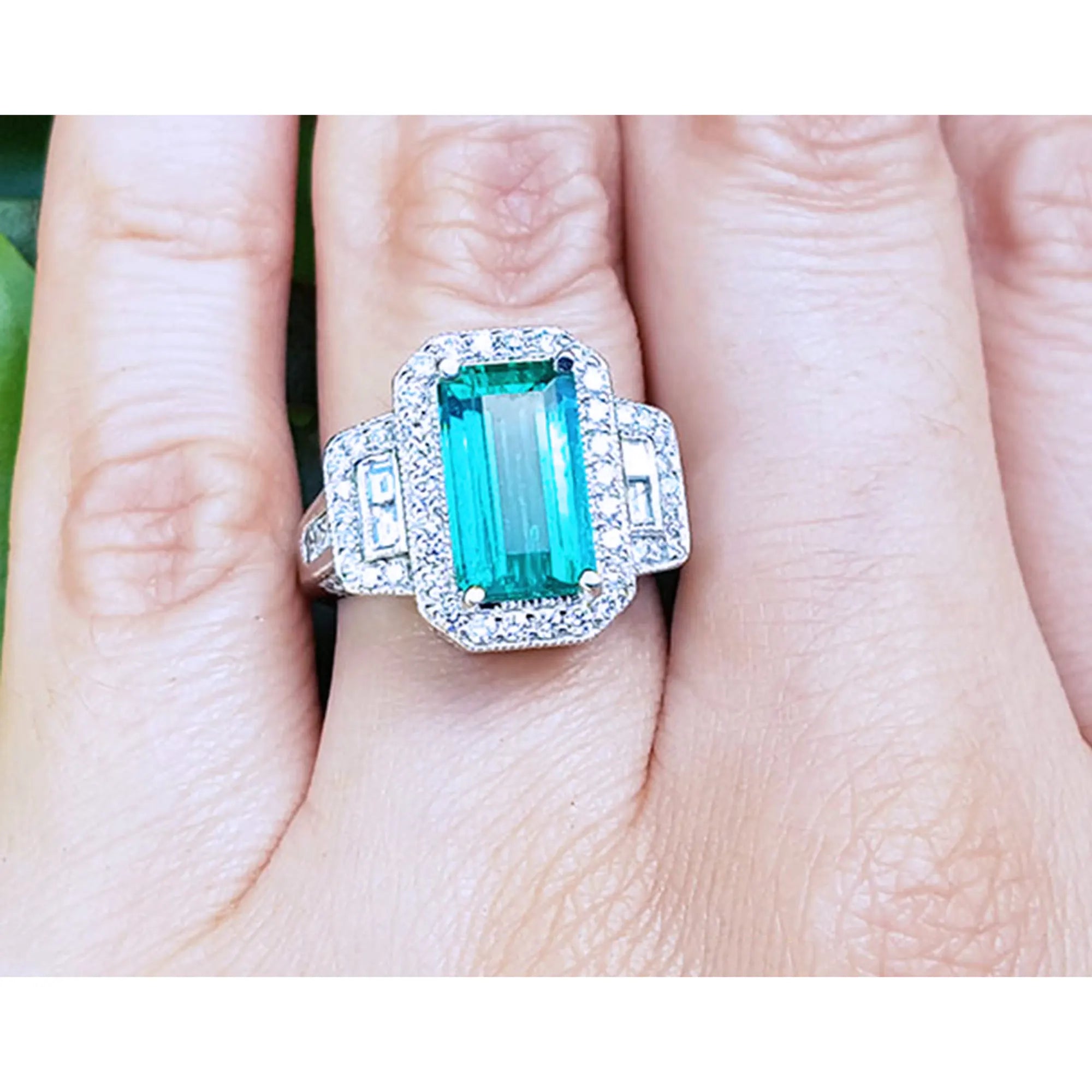 Anillo con esmeralda colombiana rectangular rodeada de diamantes en oro blanco exhibida entre dedos.