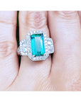 Anillo con esmeralda colombiana rectangular rodeada de diamantes en oro blanco exhibida entre dedos.