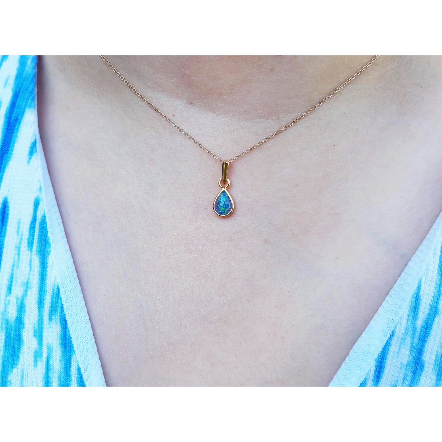 Teardrop-shaped blue opal pendant on a gold chain worn by a person with a blue and white patterned top.