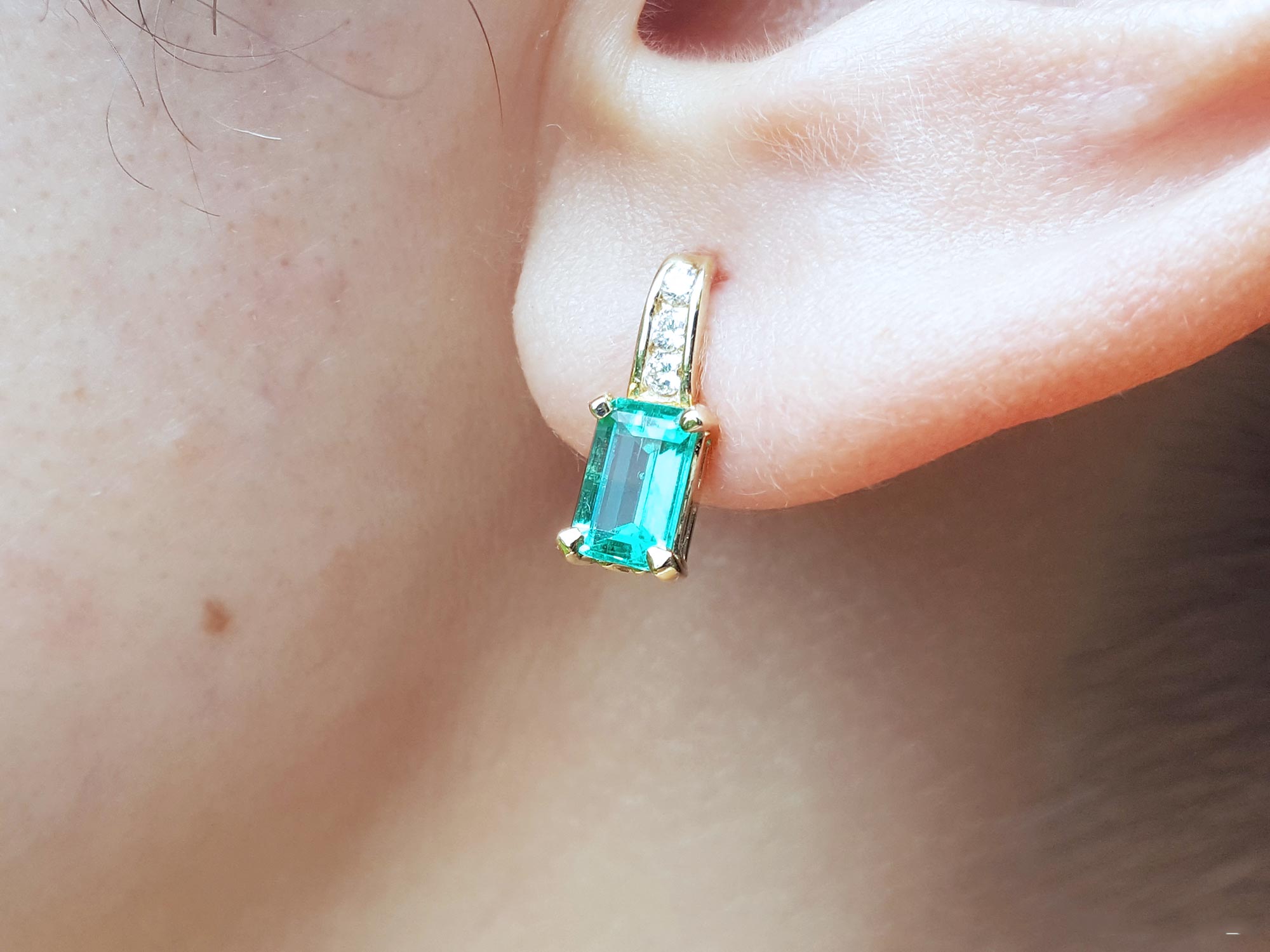 A close up of a yellow gold emerald earring, top accent diamonds in 14k yellow gold on an earlobe