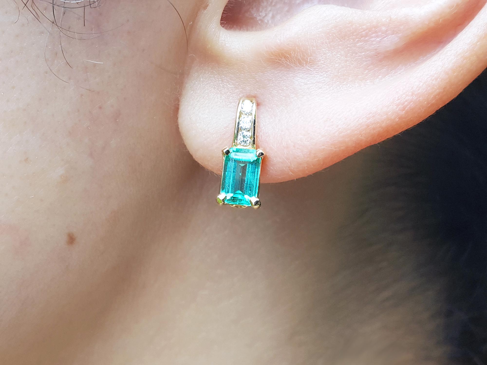 A closer front view of a yellow gold emerald earring emerald-cut with accent diamonds on top, on an woman&#39;s ear