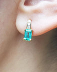 A closer front view of a yellow gold emerald earring emerald-cut with accent diamonds on top, on an woman's ear