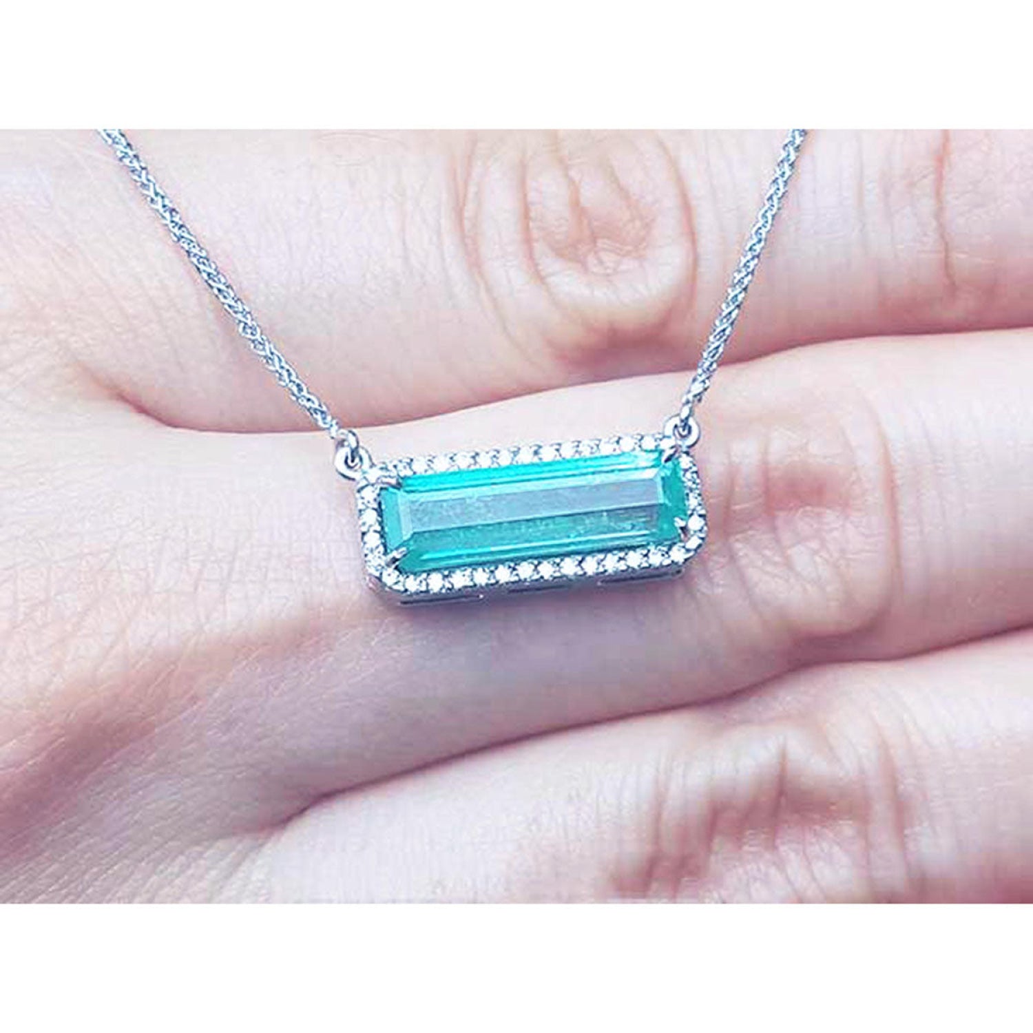 18K white gold necklace with a rectangular Colombian emerald and diamond halo, East-West design, shown resting on a person's fingers.