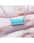 18K white gold necklace with a rectangular Colombian emerald and diamond halo, East-West design, shown resting on a person's fingers.
