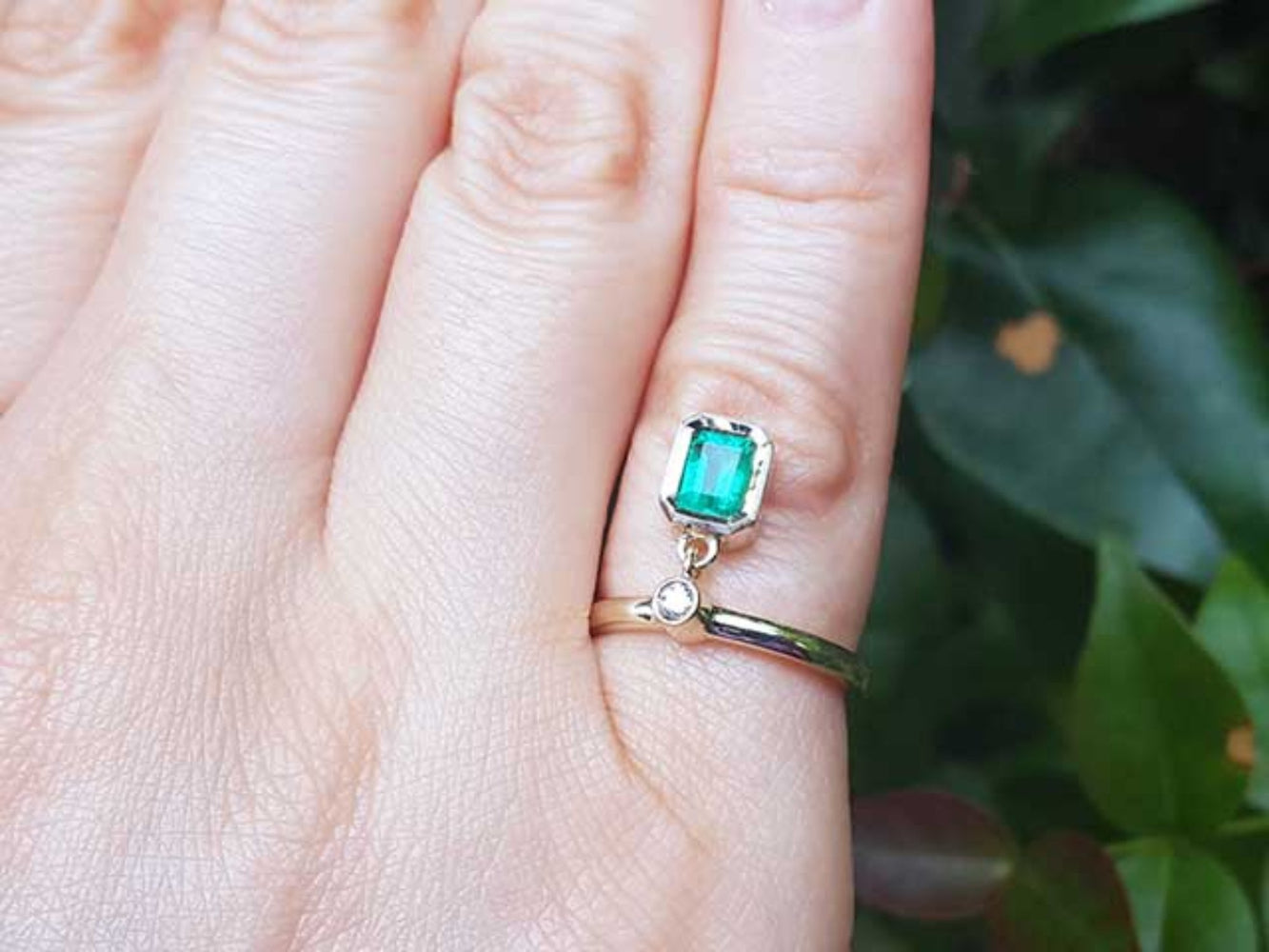 Close-up of a hand wearing emerald dangle ring May&#39;s birthstone jewelry against a blurred green background.