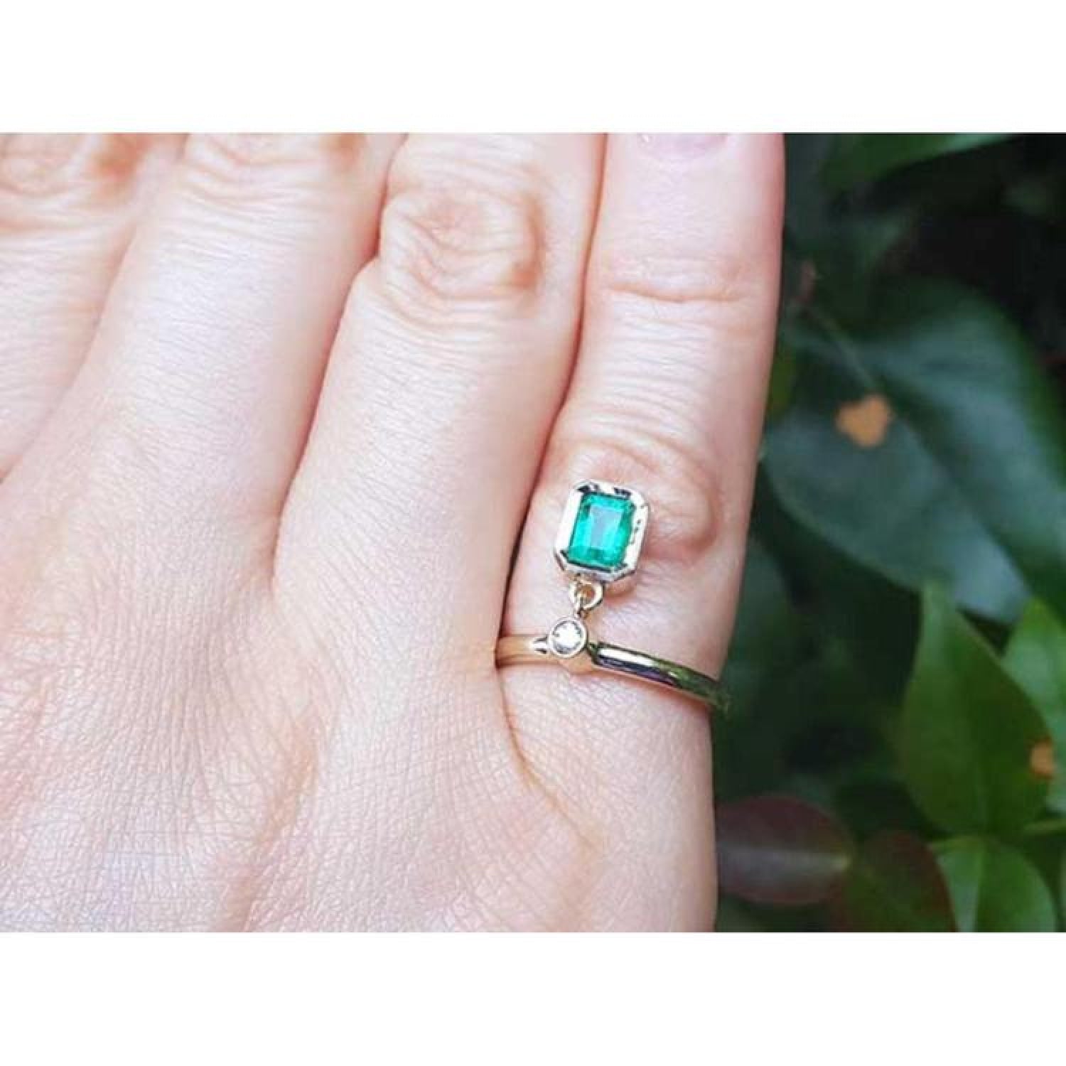 14K yellow gold dangle ring with a bezel-set emerald and small diamond on a finger.