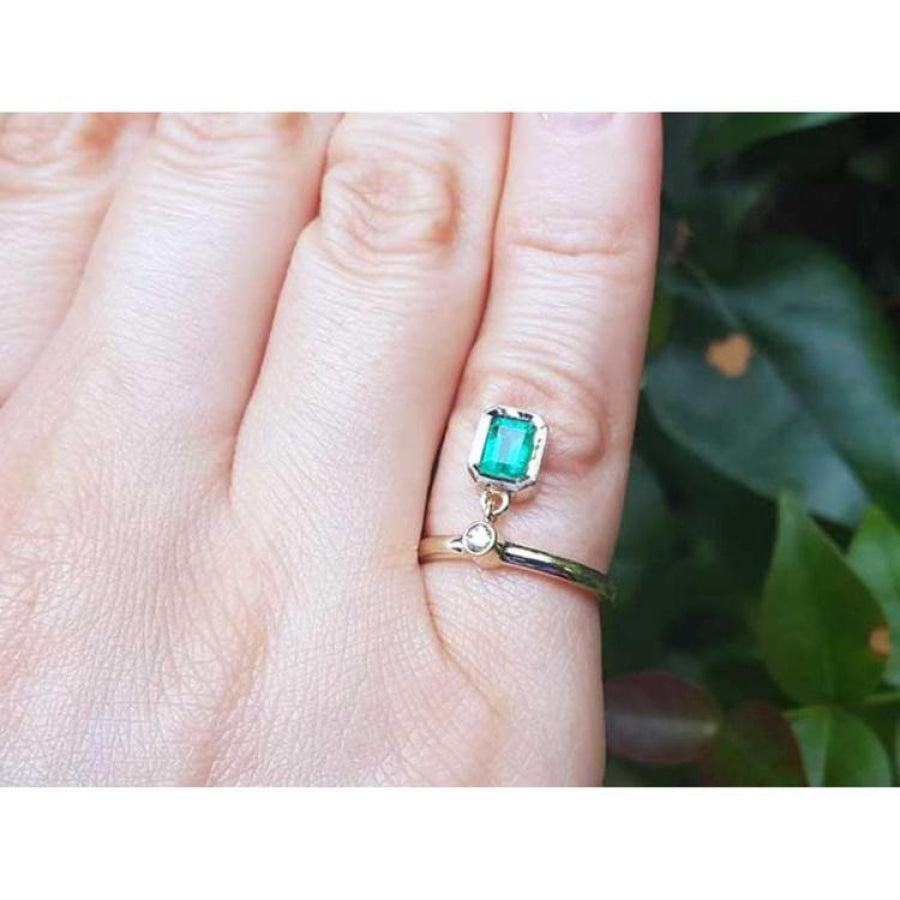 14K yellow gold dangle ring with a bezel-set emerald and small diamond on a finger.