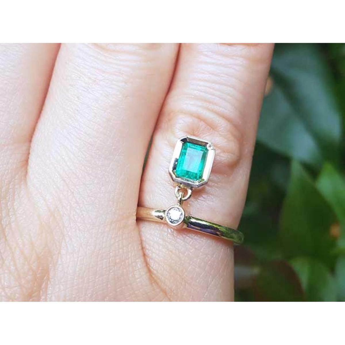 A yellow gold emerald dangle ring on a finger against a blurred natural background.