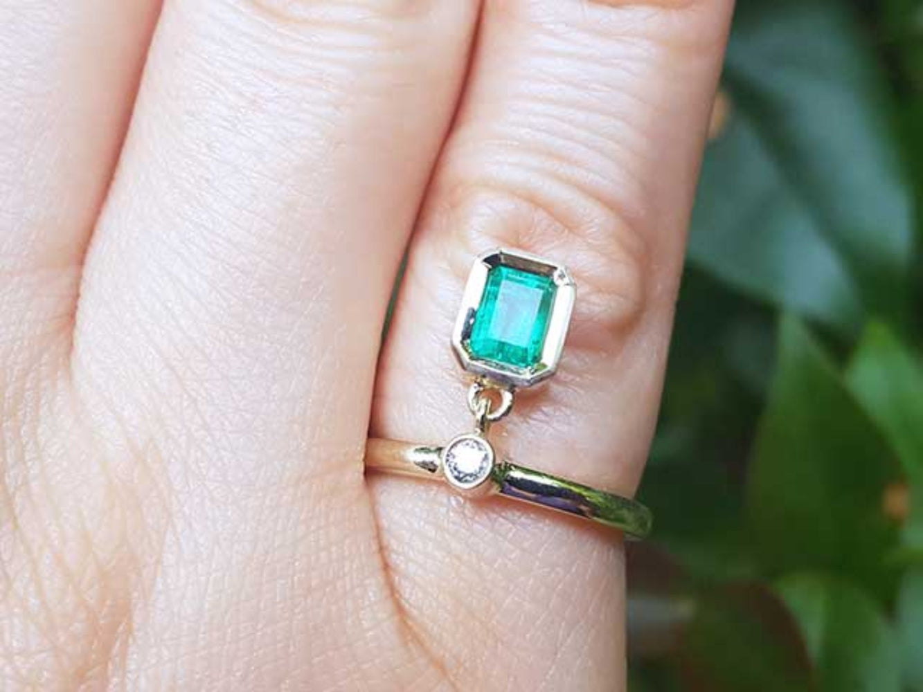 A yellow gold emerald dangle ring on a finger against a blurred natural background.