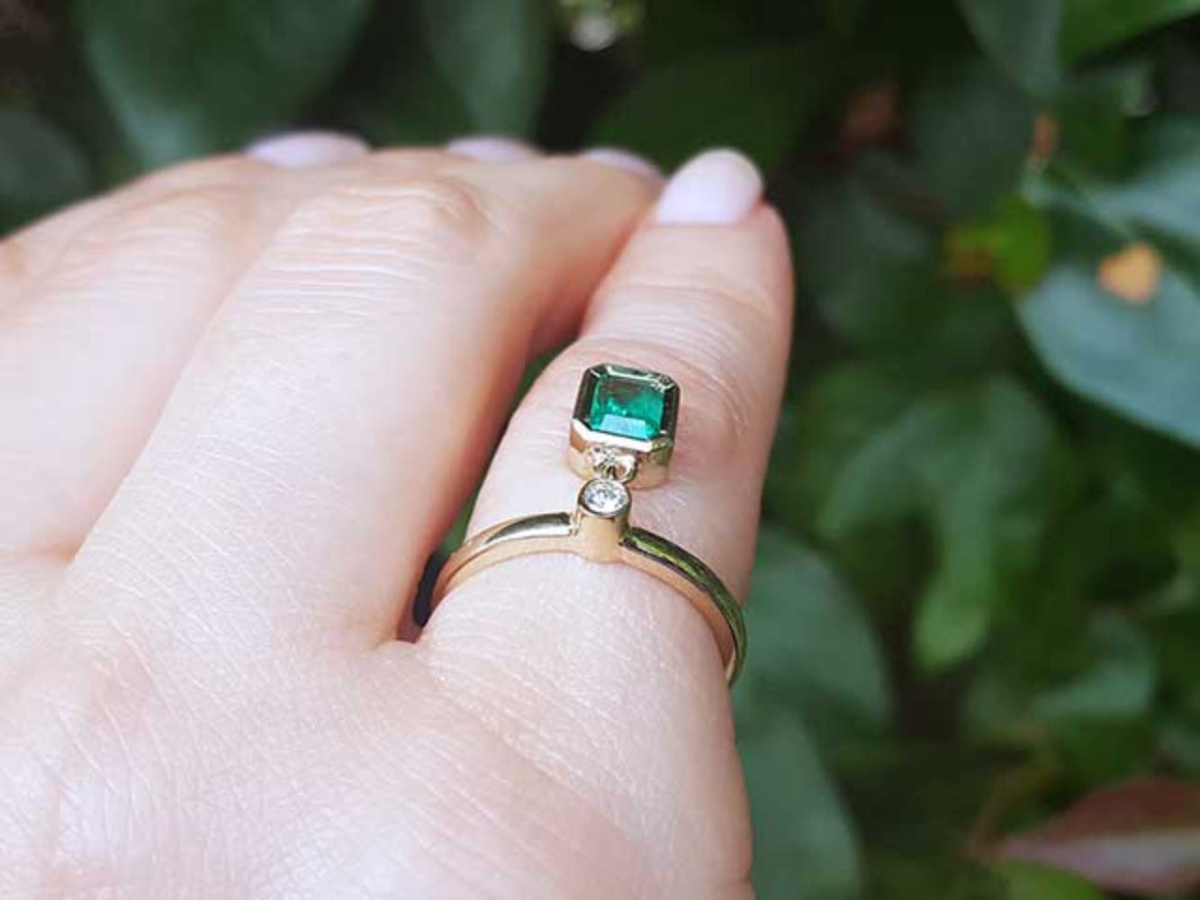 An emerald dangle ring representing May&#39;s birthstone jewelry on a hand against a blurred green leafy background.