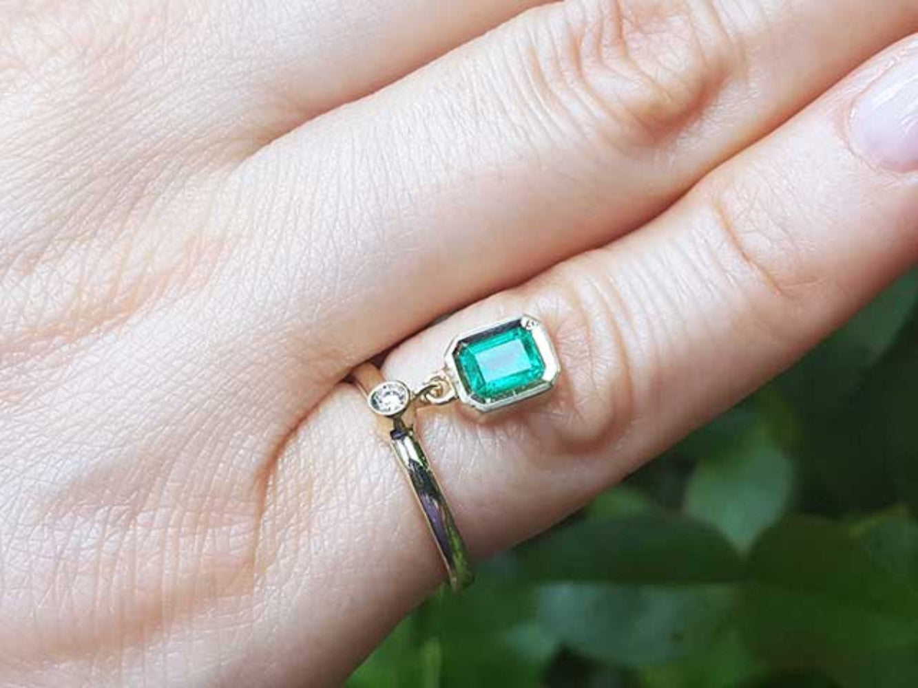 An emerald dangle ring representing May&#39;s birthstone jewelry on a hand against a blurred green background.