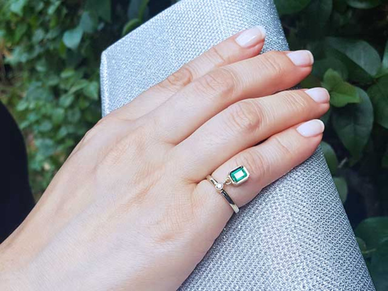 Hand wearing an emerald dangle ring against a blurred natural background.