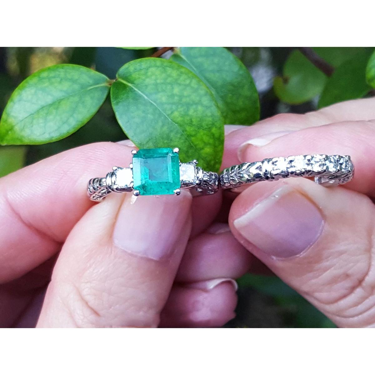 Between fingers holding an emerald ring and its matching diamond wedding band on a leafy background.