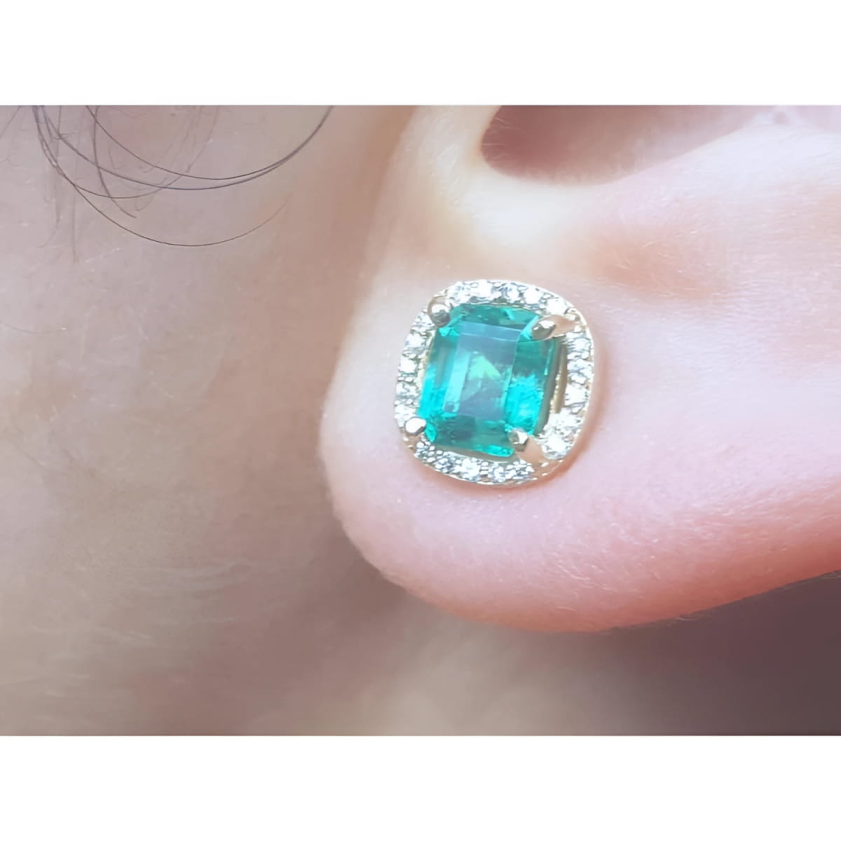 Close-up of an emerald halo diamond stud earring on a person's ear.
