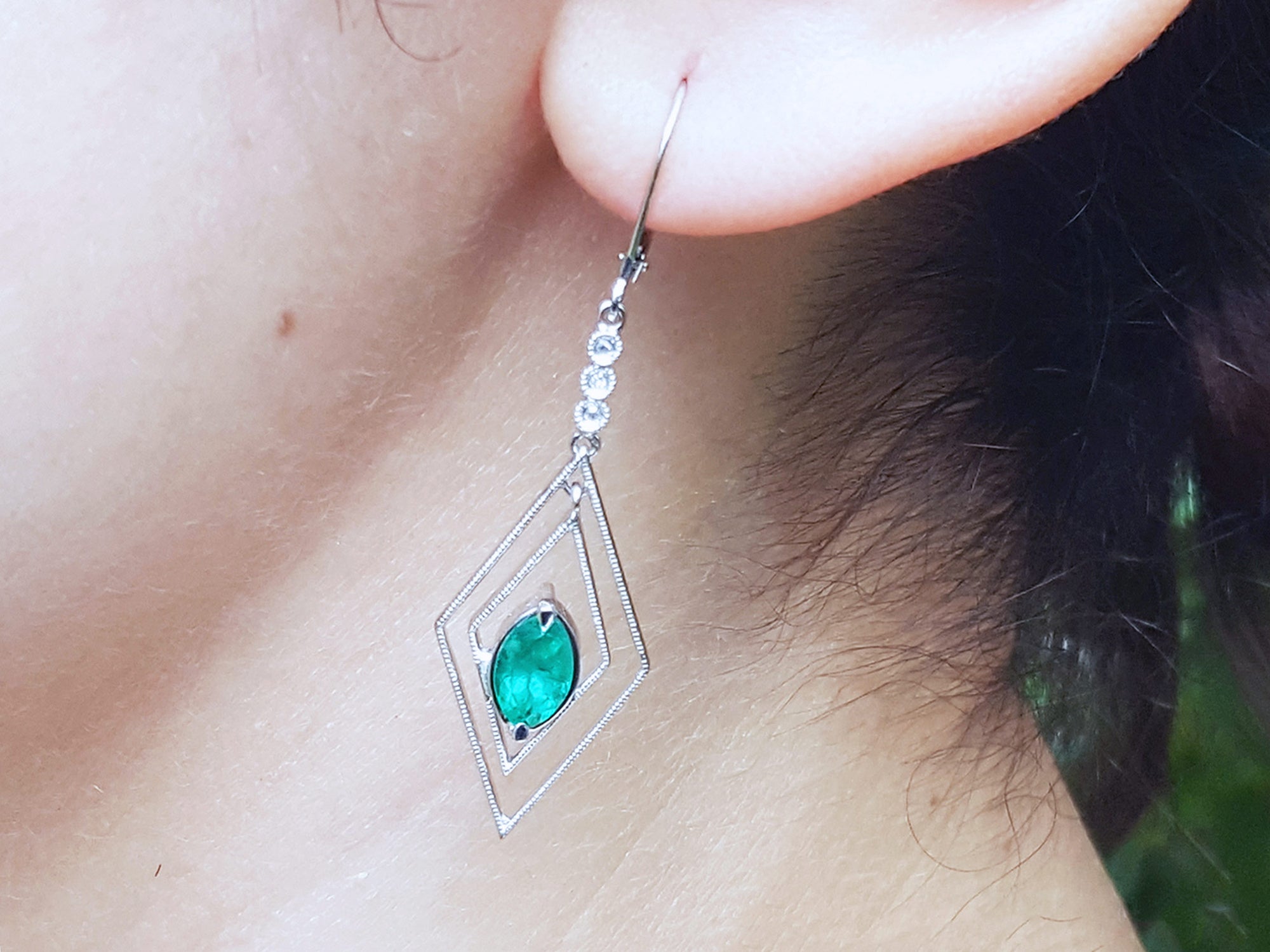 Close-up of a diamond-shaped earring with a marquise emerald worn by a woman.