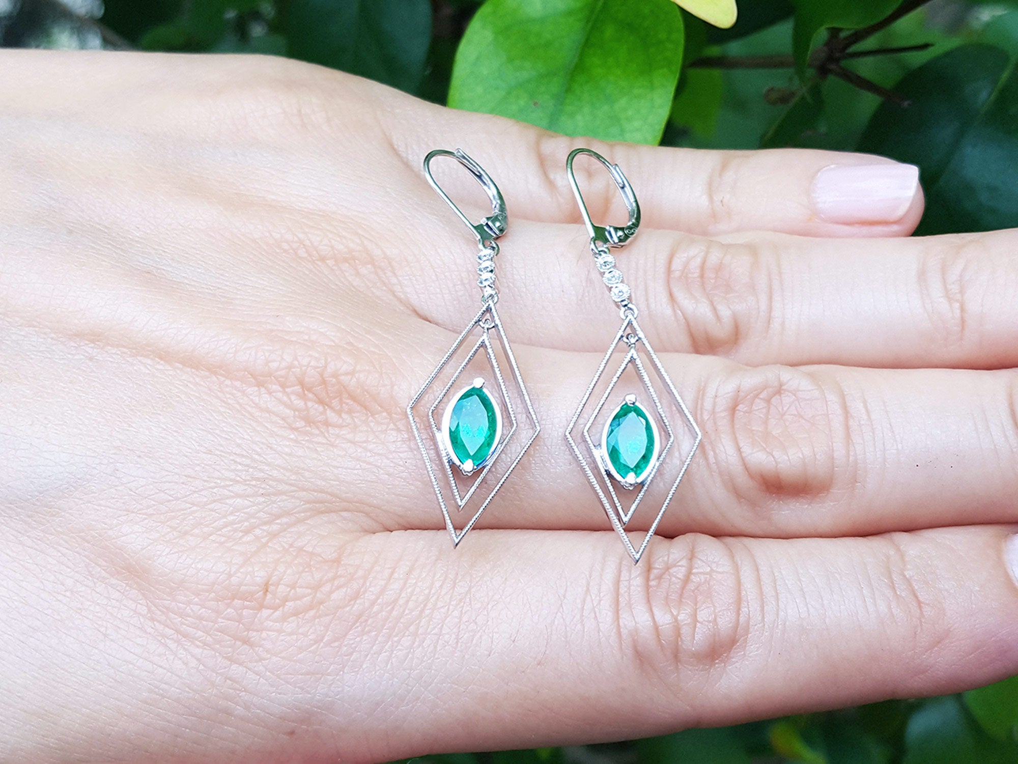 Diamond-shaped earrings with marquise emeralds gemstones placed on fingers against a natural background.