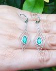 Diamond-shaped earrings with marquise emeralds gemstones placed on fingers against a natural background.