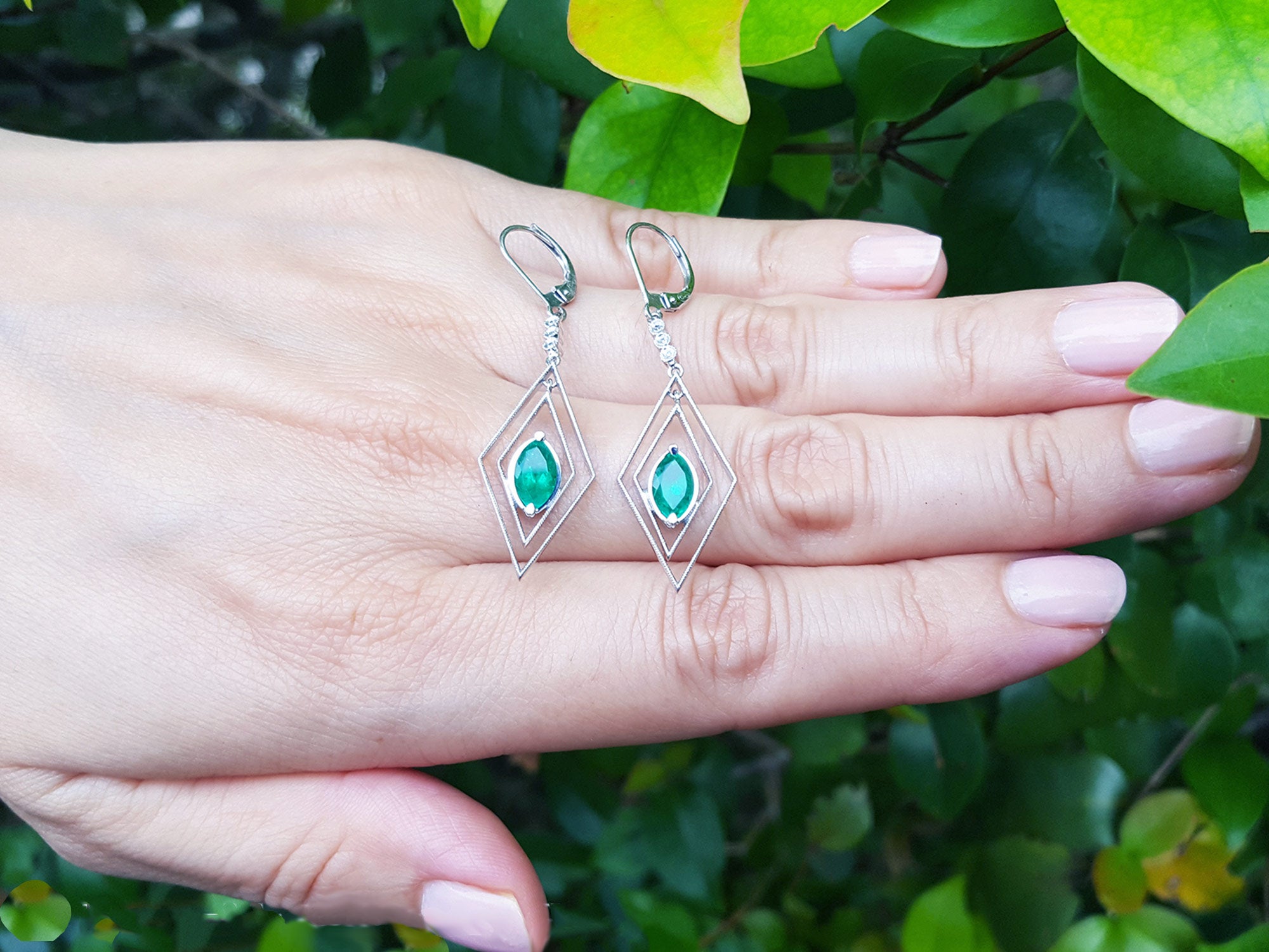 Diamond-shaped earrings with marquise emerald gemstones on a hand against a green leafy background