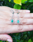 Diamond-shaped earrings with marquise emerald gemstones on a hand against a green leafy background