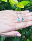 Hand displaying a pair of diamond-shaped emerald marquise earrings against a green leafy background.
