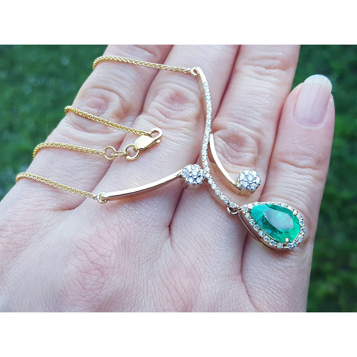 Hand holding a yellow gold necklace with a natural emerald gemstone pendant against a blurred green background