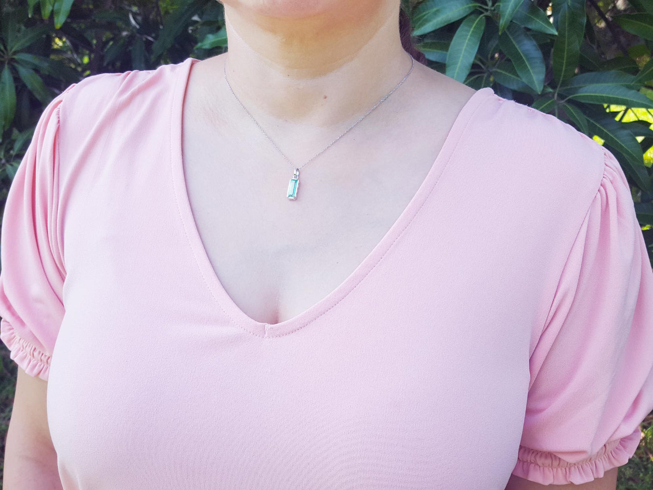 A woman wearing a pink top with a Colombian emerald pendant against a green leafy background.