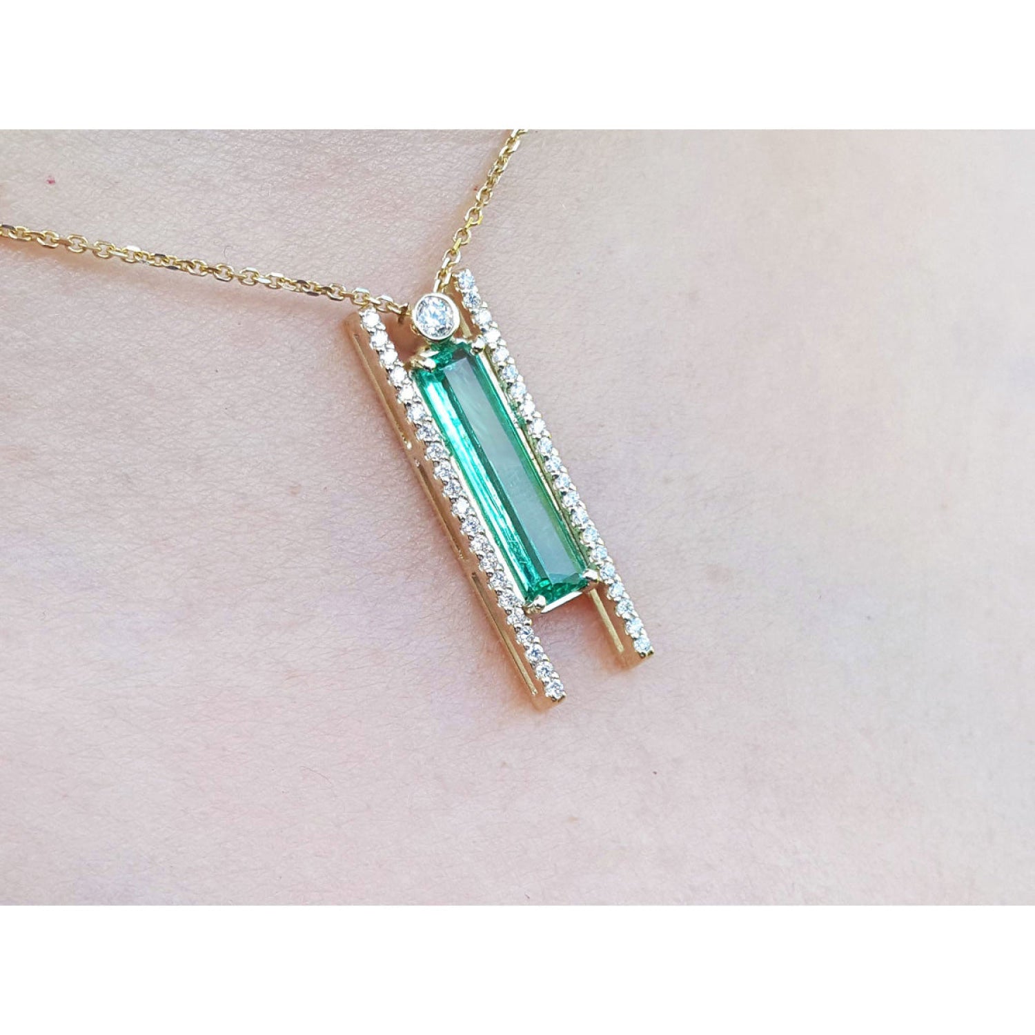 Emerald baguette cut necklace with natural diamonds, 14K yellow gold, and sliding tube bail chain., shown worn on a person's neck against a pale skin background.