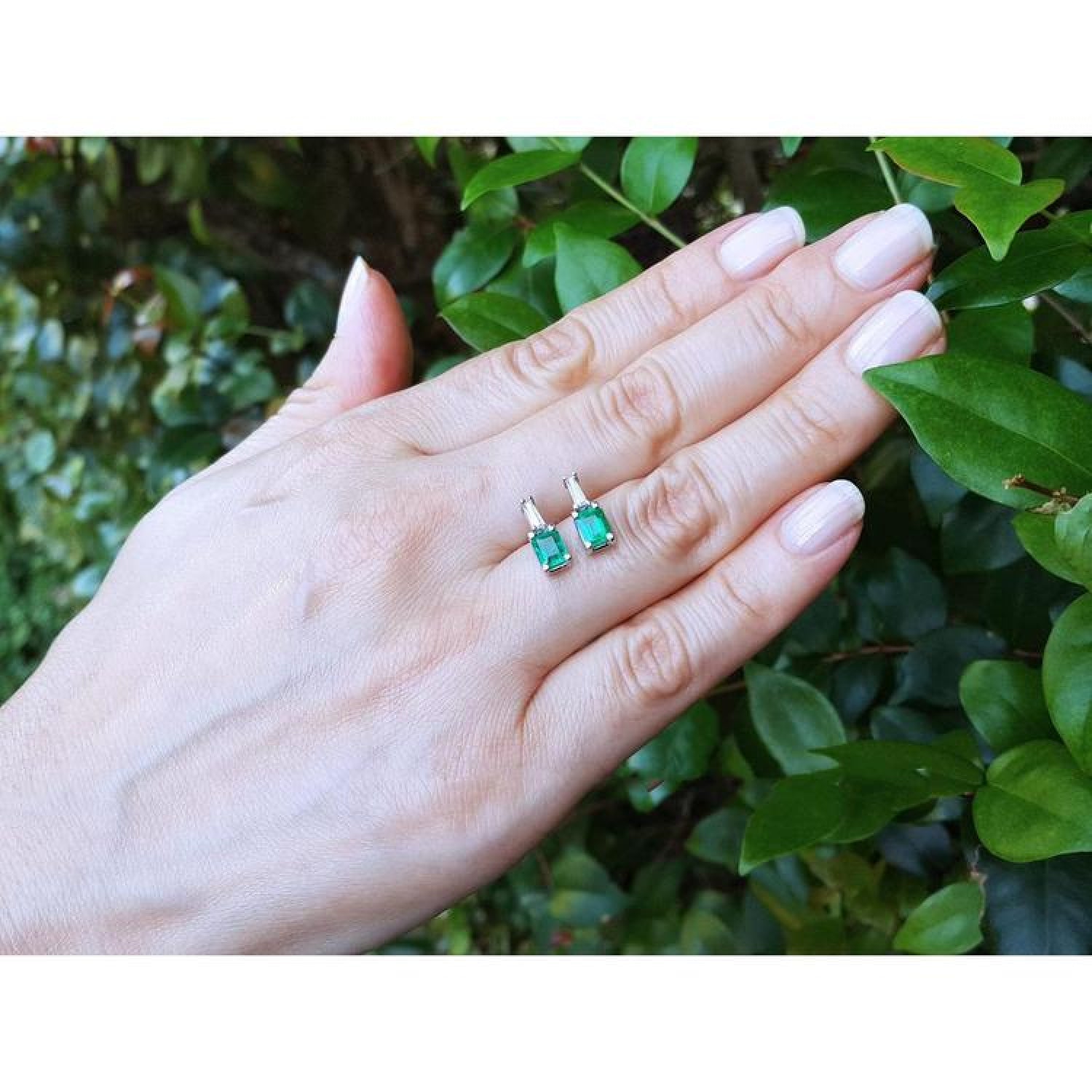 Colombian emerald and baguette diamond stud earrings, 14K gold, on a hand with green leaves.