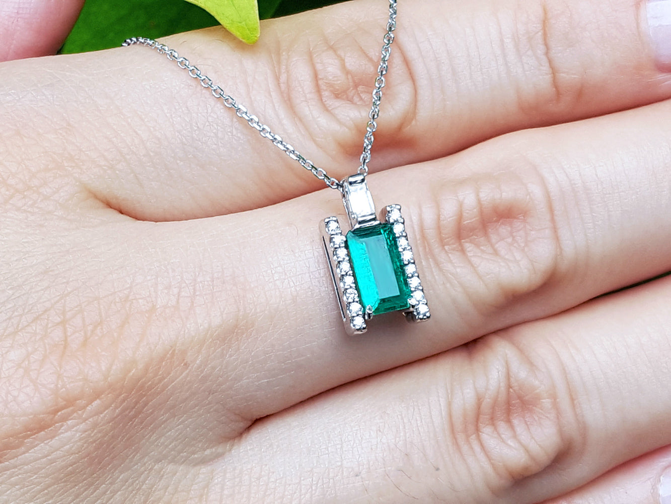 Baguette cut emerald gemstone pendant on a white gold chain held between fingers.