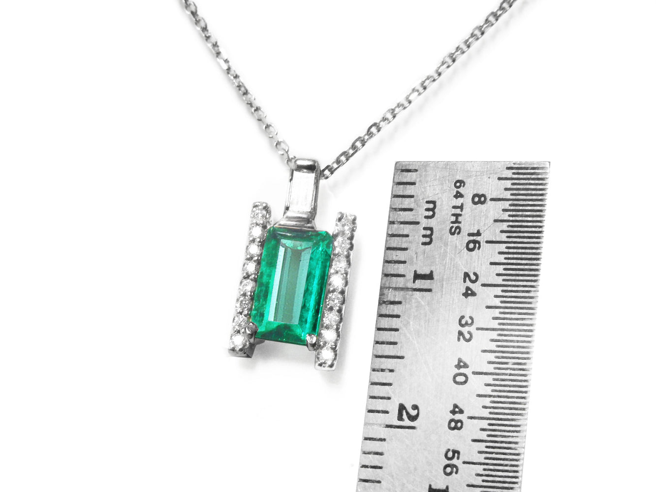 Emerald and diamond baguette pendant on a chain with a ruler for scale on a white background.