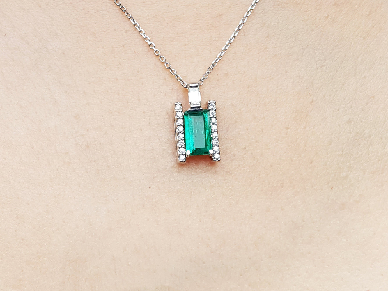 Emerald gemstone pendant on a white gold chain against a beige skin background.