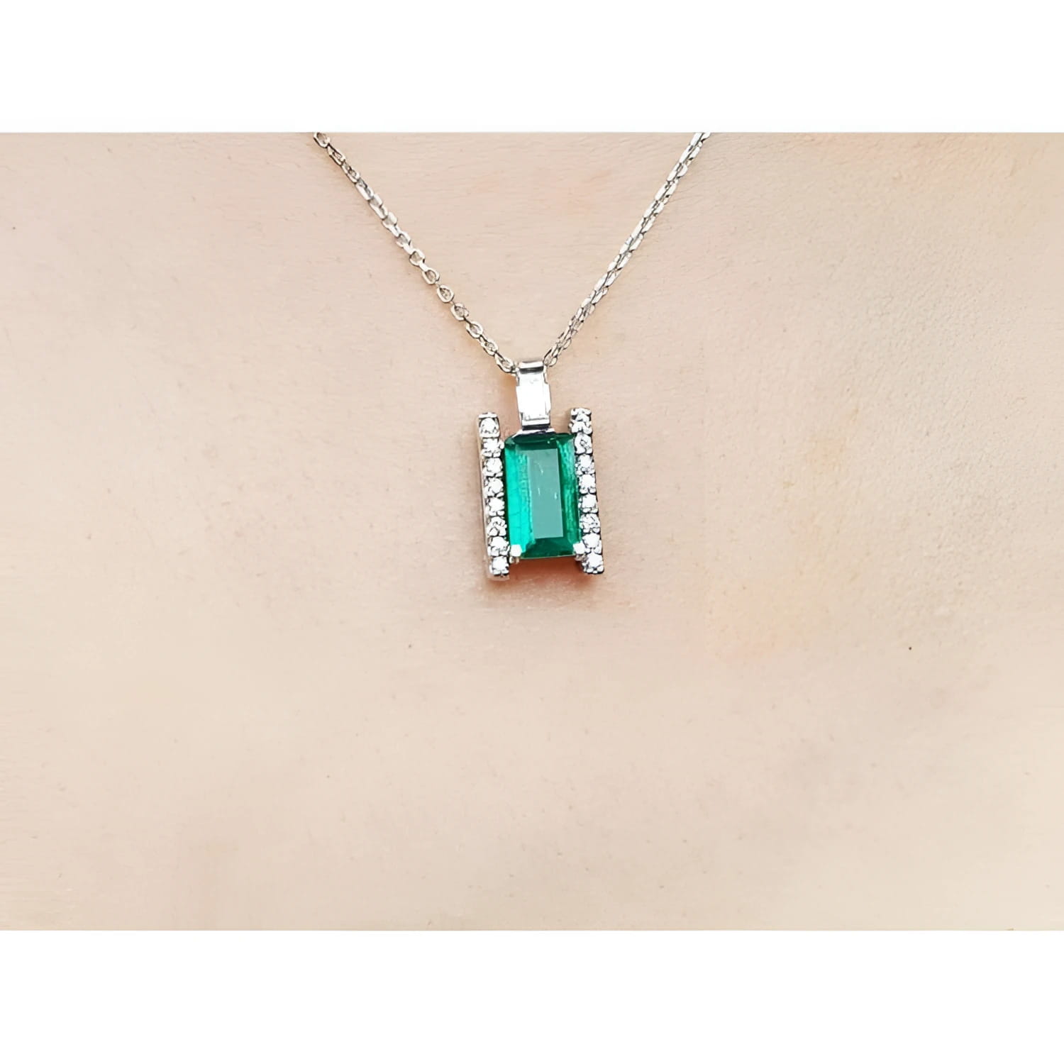 Emerald gemstone pendant on a white gold chain against a beige skin background.