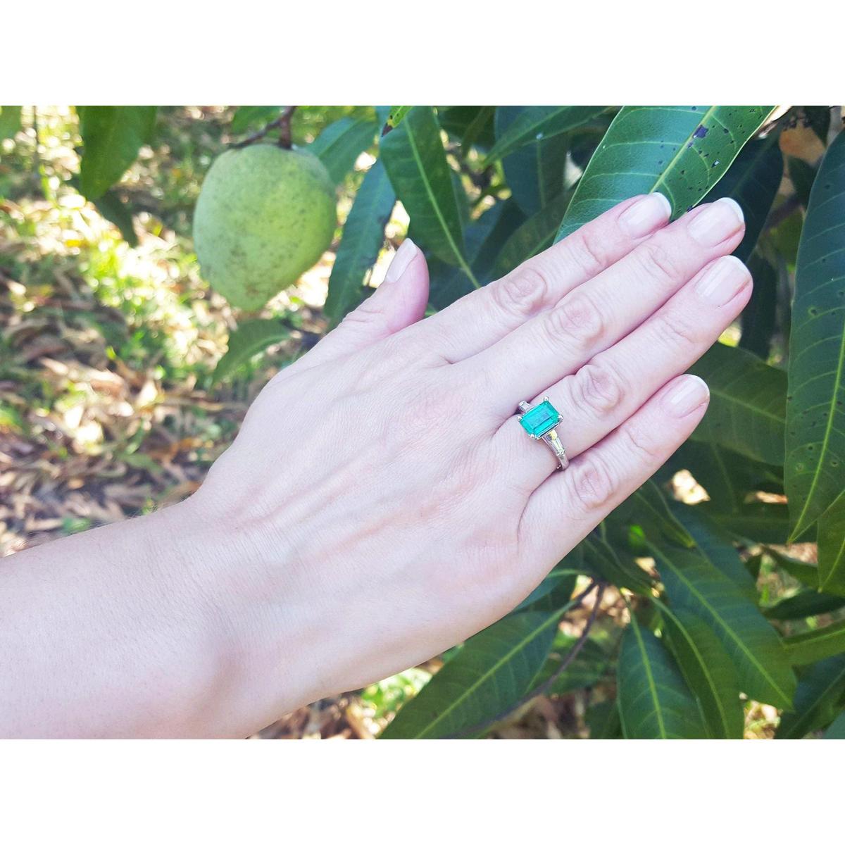 Hand with a three stone emerald and baguette side diamond ring in front of green leaves.