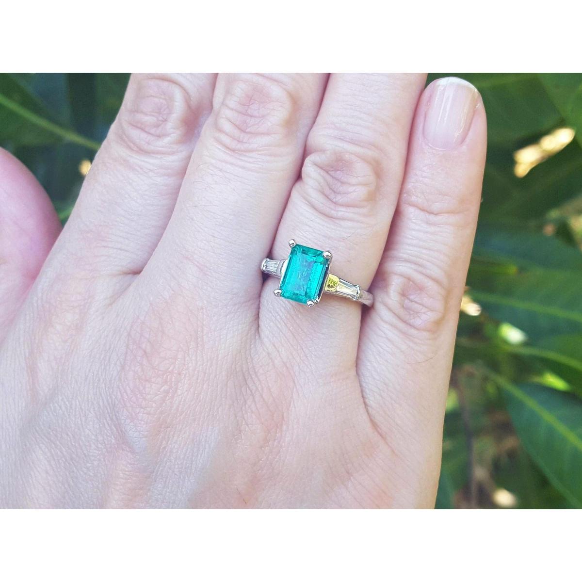 Ring with an emerald and baguette side diamond ring on a person's finger against a blurred green leafy background.