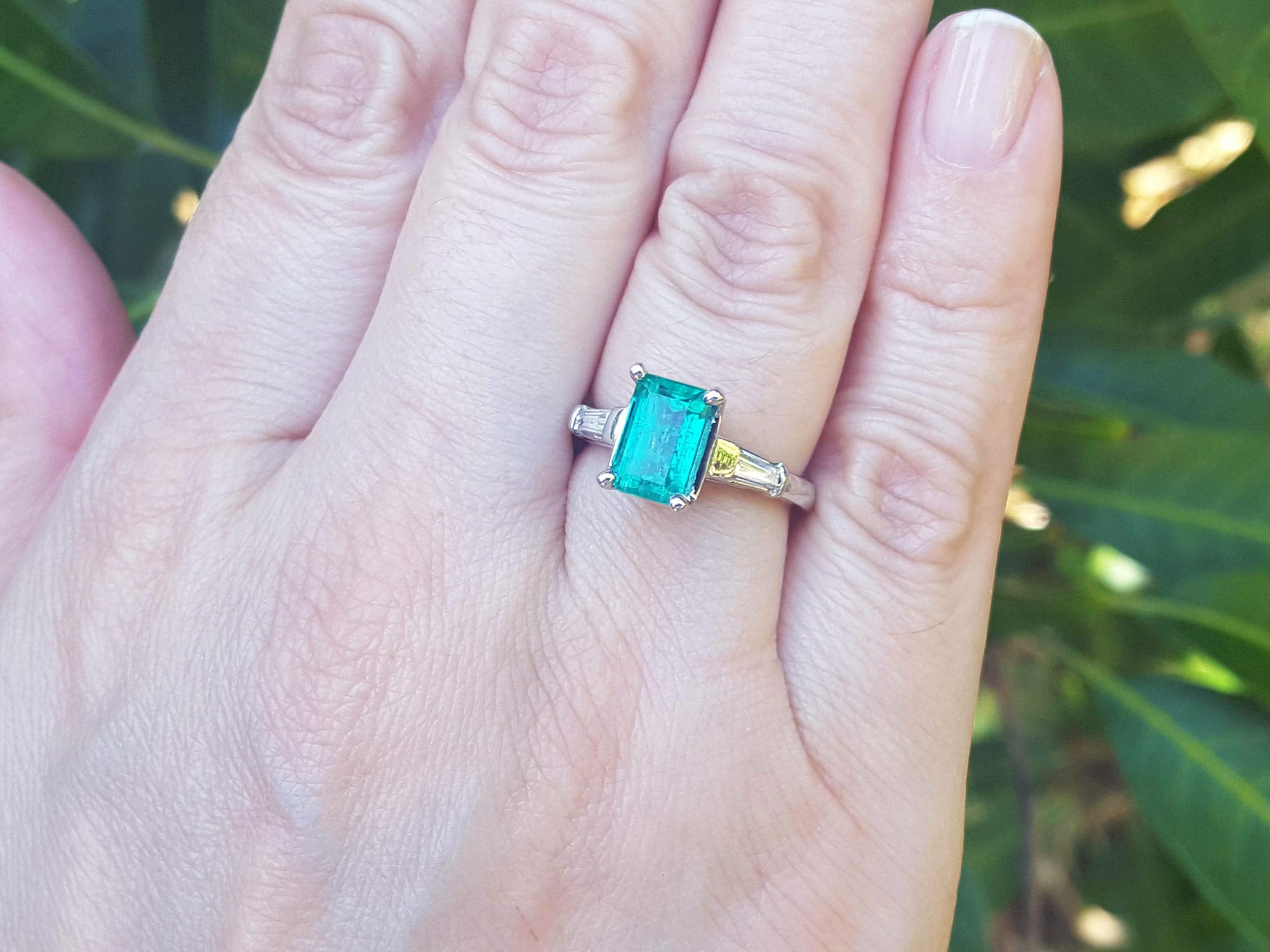 Ring with an emerald and baguette side diamond ring on a person&#39;s finger against a blurred green leafy background.