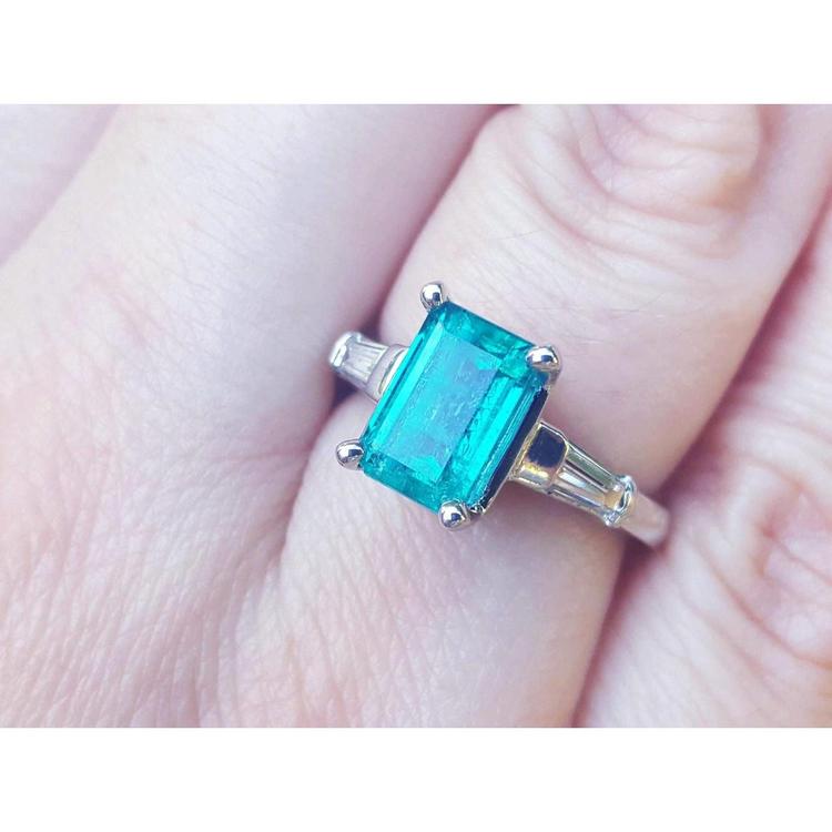 A three stone genuine emerald and baguette side diamond ring in white gold on a finger.