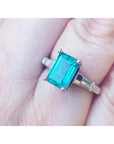 A three stone genuine emerald and baguette side diamond ring in white gold on a finger.