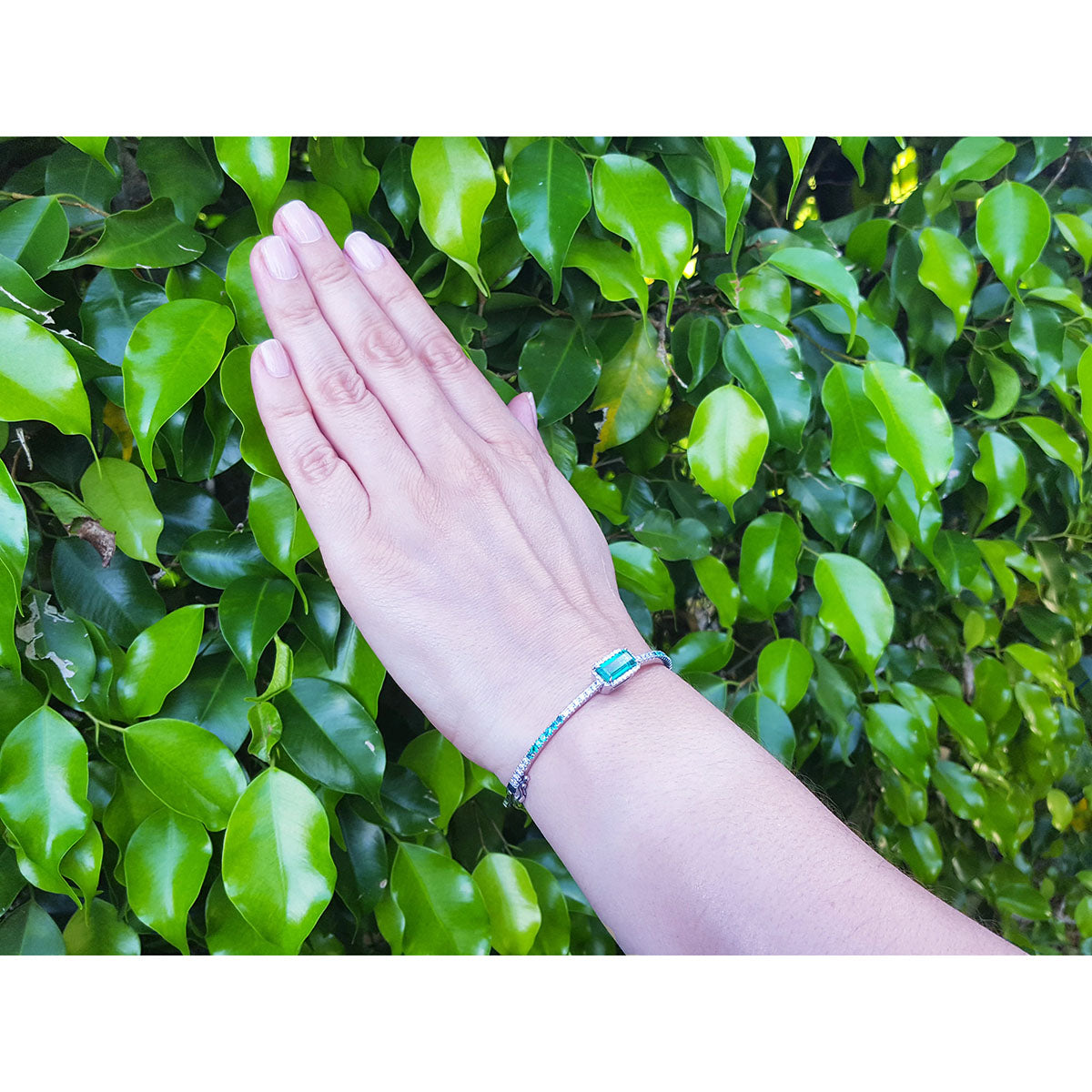 A woman&#39;s hand wearing a bangle bracelet with emeralds and diamonds against a green leafy background.