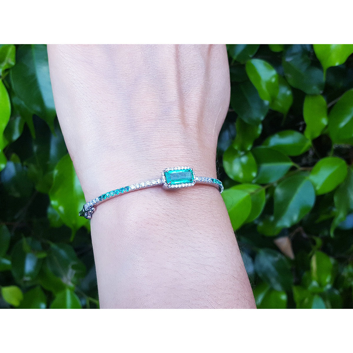 A bangle bracelet with a rectangular emerald and diamonds on a wrist against a green leafy background.