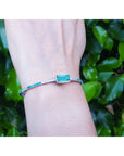 A bangle bracelet with a rectangular emerald and diamonds on a wrist against a green leafy background.