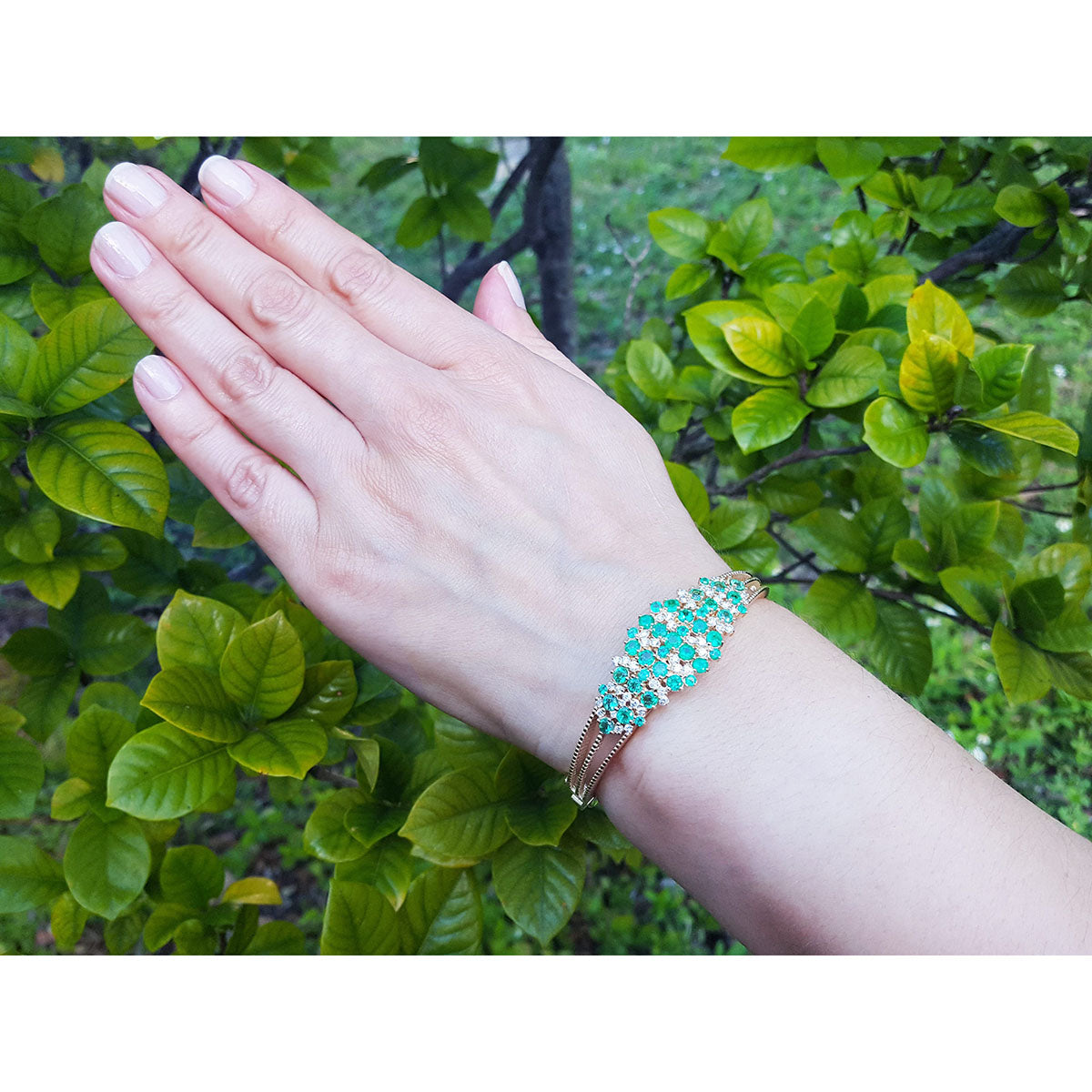 Hand wearing a natural emeralds and diamonds bangle bracelet with a green leafy background.